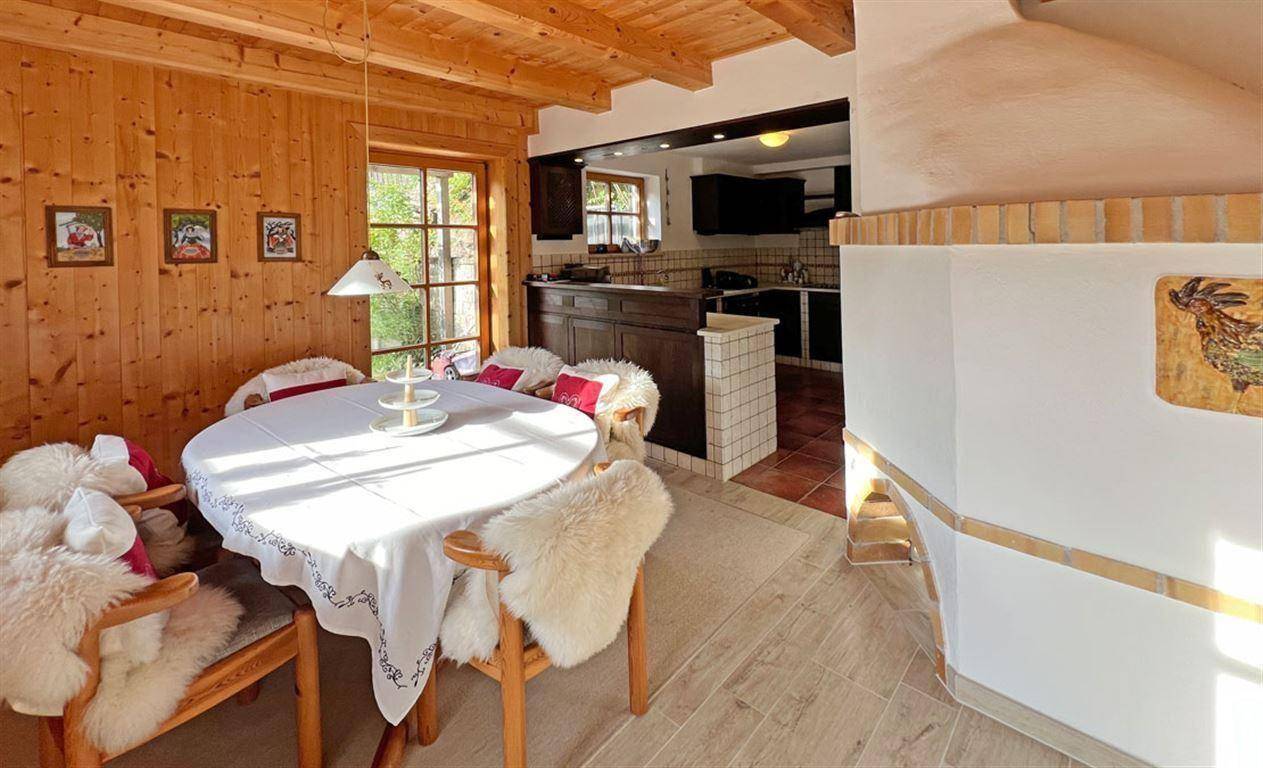 Offener Essbereich mit Kachelofen, der in eine Küche mit dunklen Holzschränken und Terrakottafliesen übergeht.