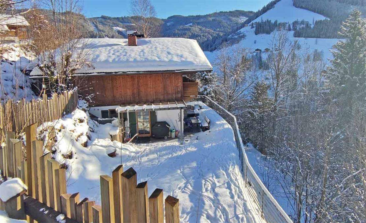 Rückseite des Chalets im Winter, umgeben von Schnee und einem Holzzaun, mit Blick auf die Skipiste.