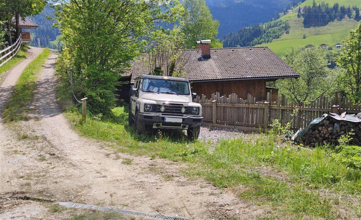 Unbefestigte Zufahrt zu einem rustikalen Holzhaus in grüner Hügellandschaft mit geparktem Geländewagen.