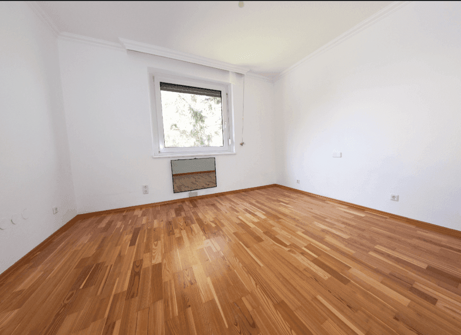 Helles, leeres Zimmer mit neuwertigem Holzparkettboden und einem Fenster mit Blick ins Grüne.
