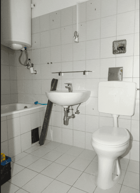 Einfaches Badezimmer mit weißen Fliesen, Badewanne, Waschbecken und Toilette, sowie einem Warmwasserboiler.