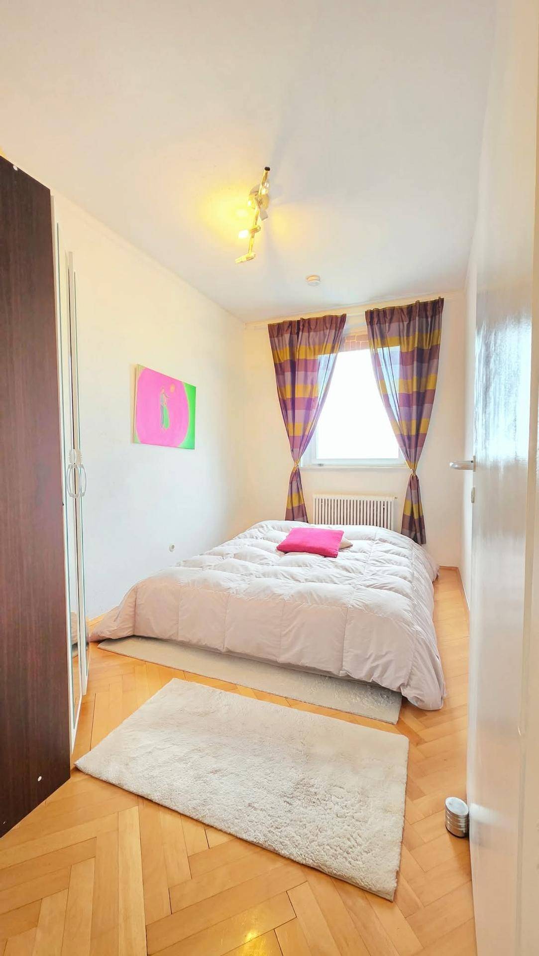 Helles Schlafzimmer mit Holzboden, einem großen Bett und farbenfrohen Vorhängen am Fenster.