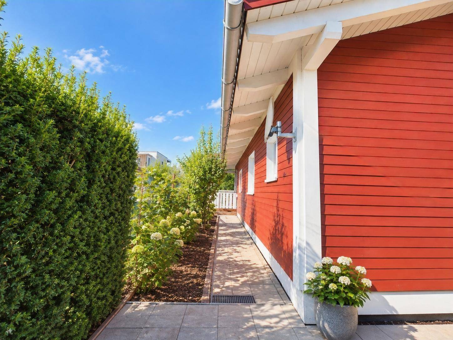 Schmaler gepflasterter Seitenweg entlang der roten Hausfassade mit hoher grüner Hecke.