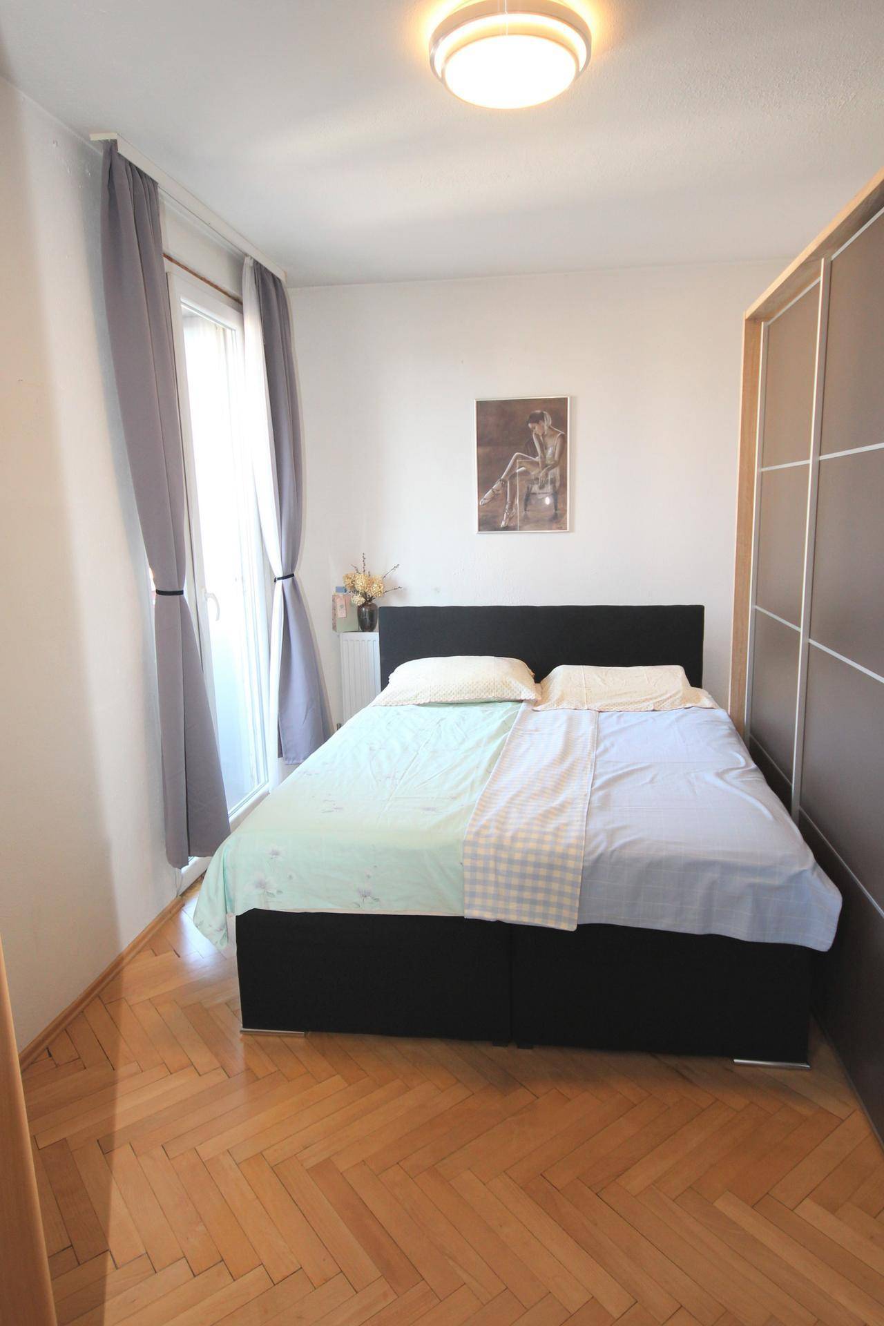 Schlafzimmer mit Doppelbett, Parkettboden und großem Fenster mit Vorhängen.