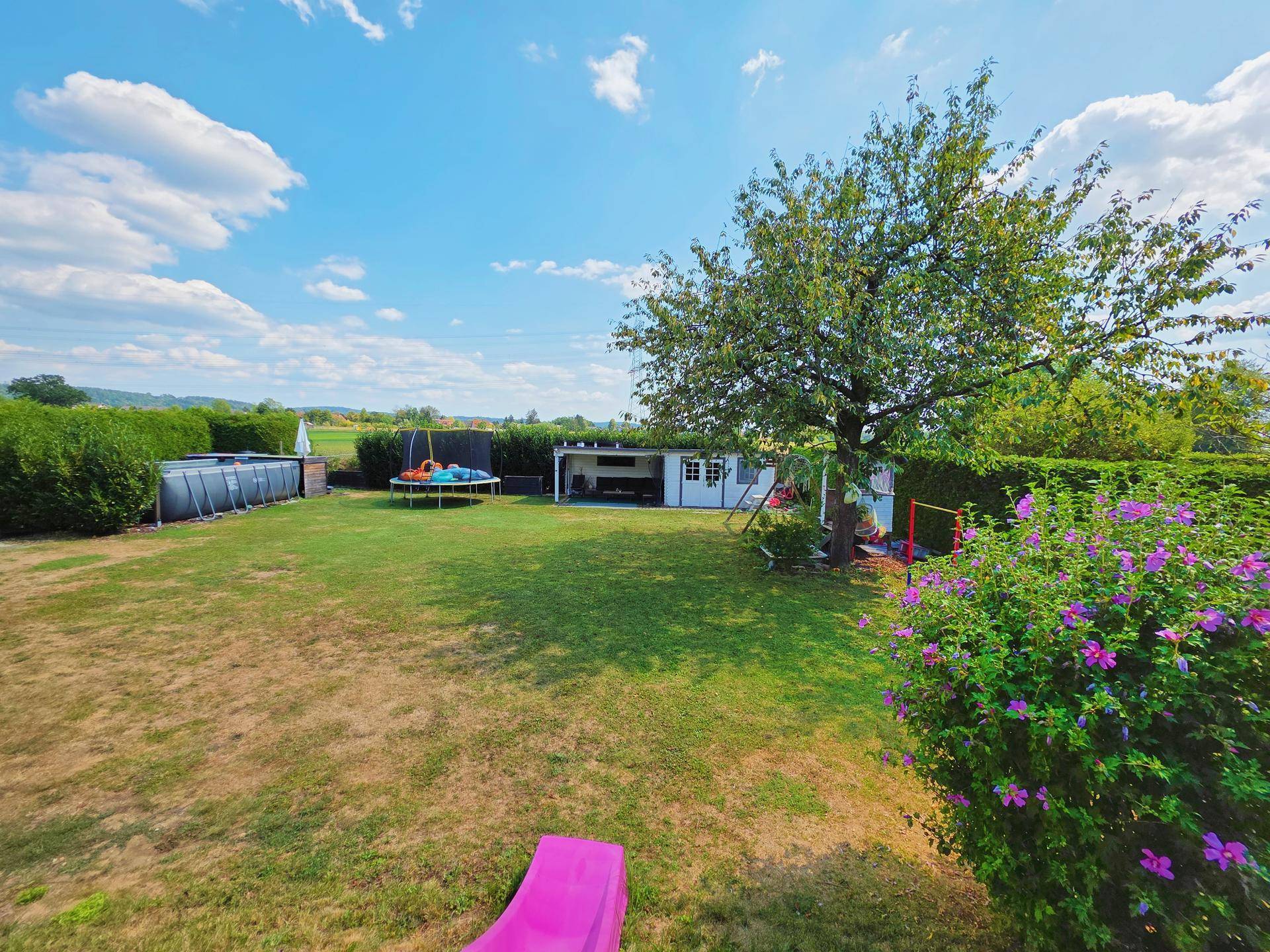Großer Garten mit Rasenfläche, Bäumen, Trampolin und Aufstellpool.