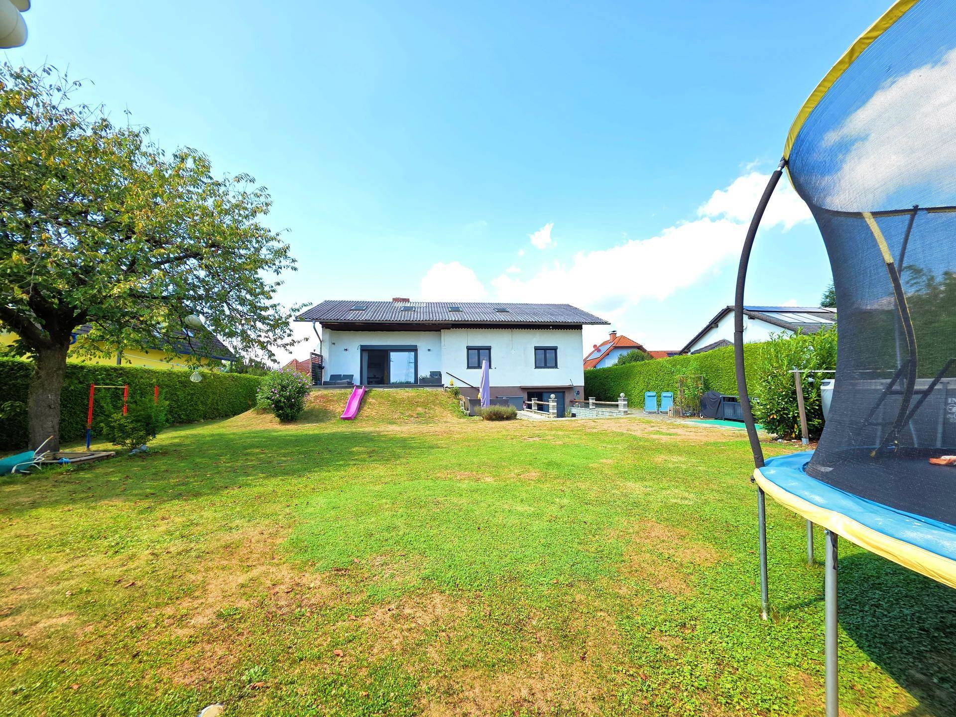 Weitläufiger Garten mit Rasen, einem Trampolin und dem Haus im Hintergrund unter blauem Himmel.