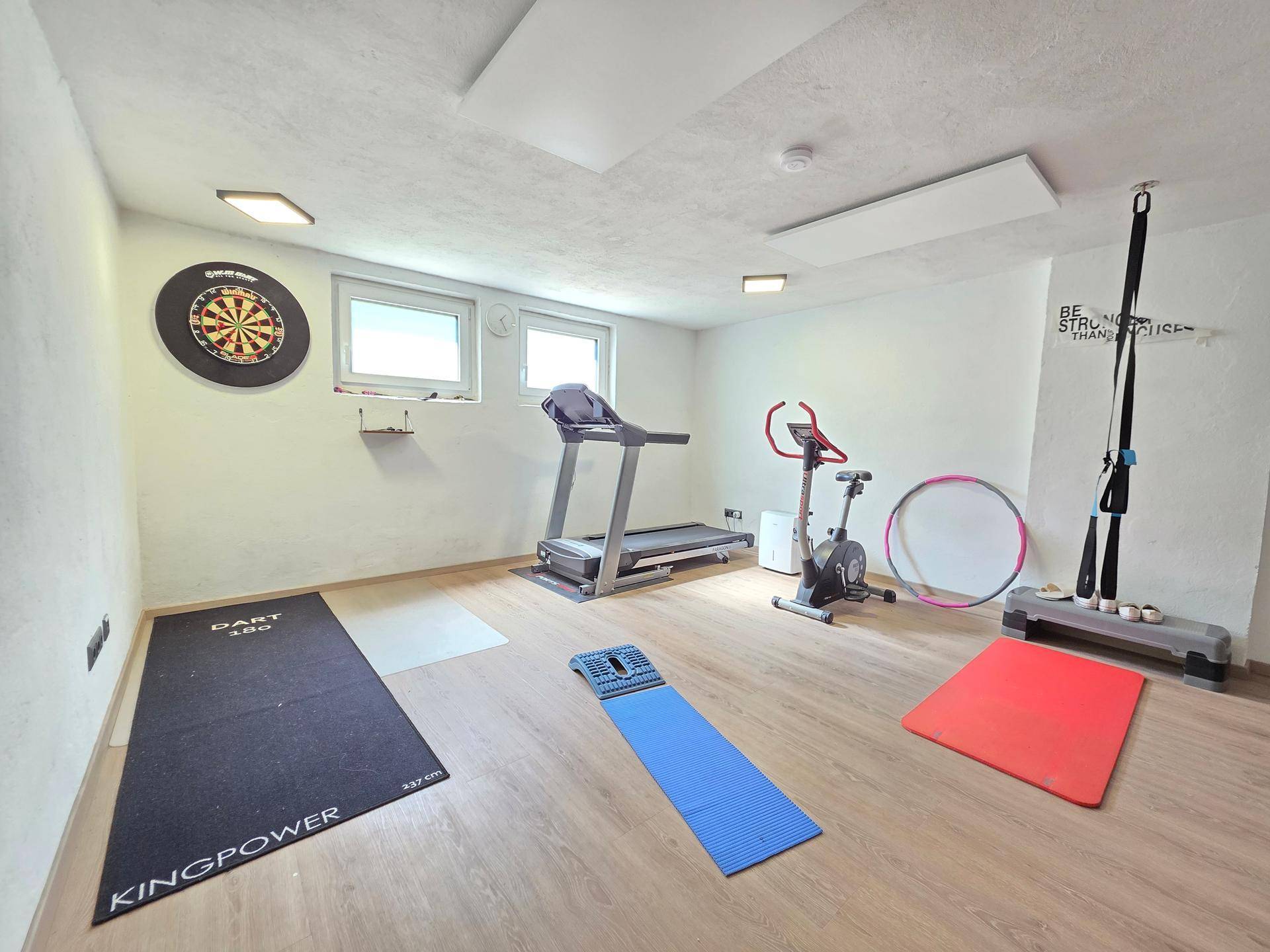 Fitnessraum im Kellergeschoss mit Laufband, Heimtrainer und Dartscheibe.