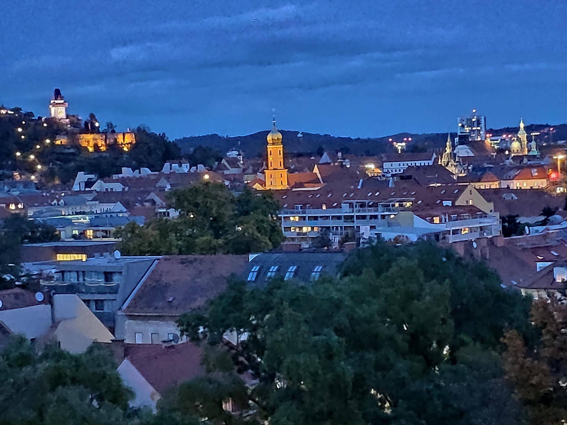 Panoramablick auf die beleuchtete Stadt bei Dämmerung mit historischen Gebäuden und Hügeln.