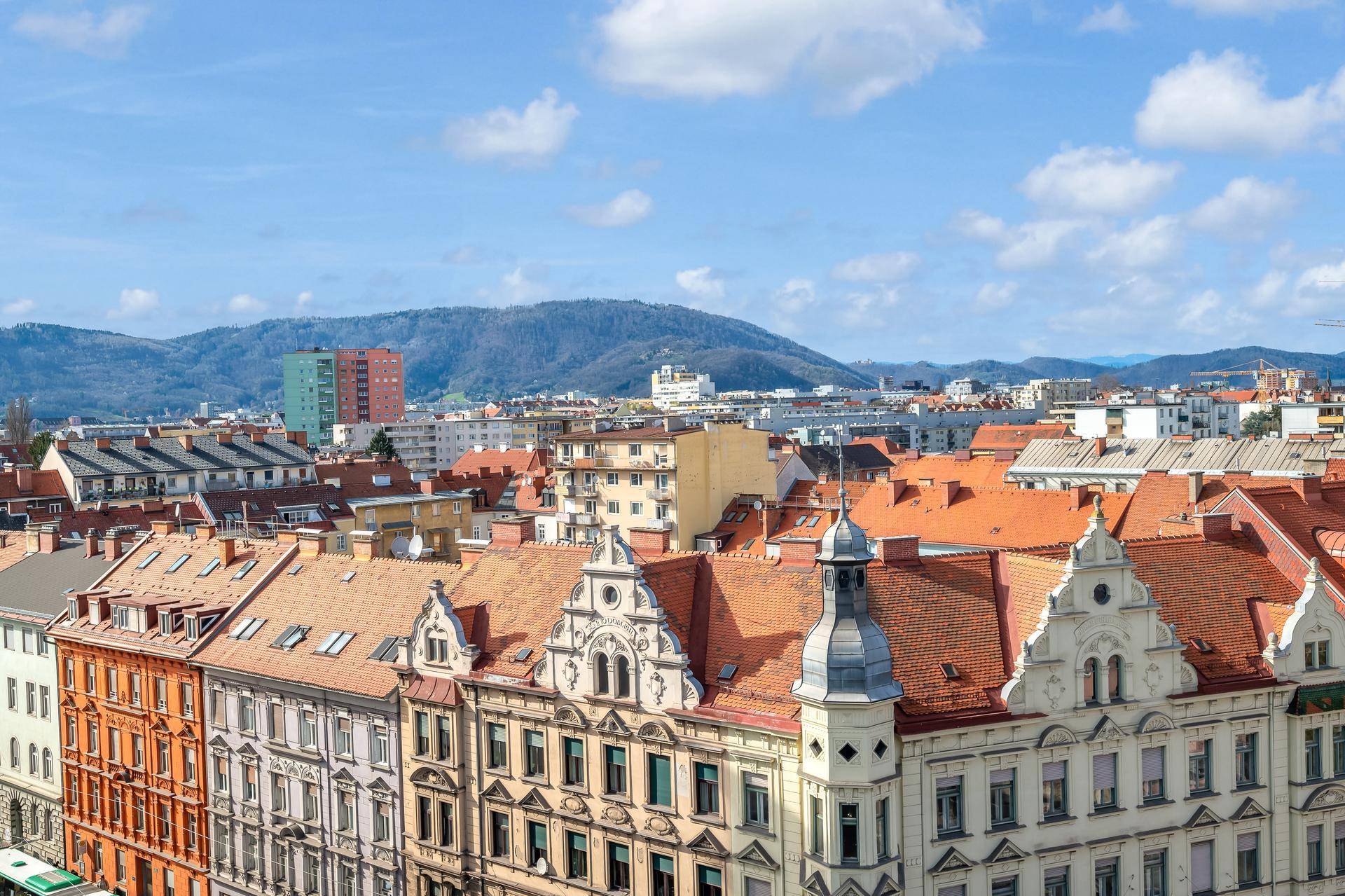 Historische Gebäude mit roten Dächern und Bergen im Hintergrund unter blauem Himmel.