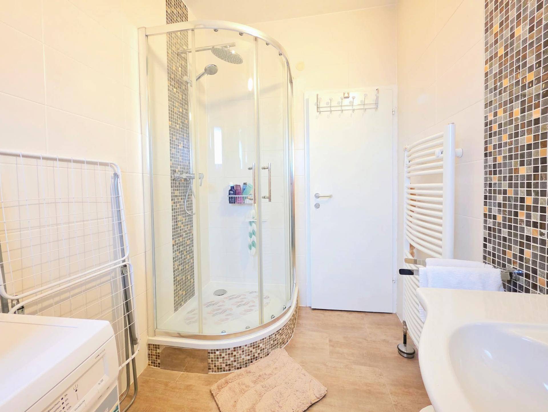 Funktionales Badezimmer mit Eckdusche, Waschbecken, beheiztem Handtuchhalter und Waschmaschine.