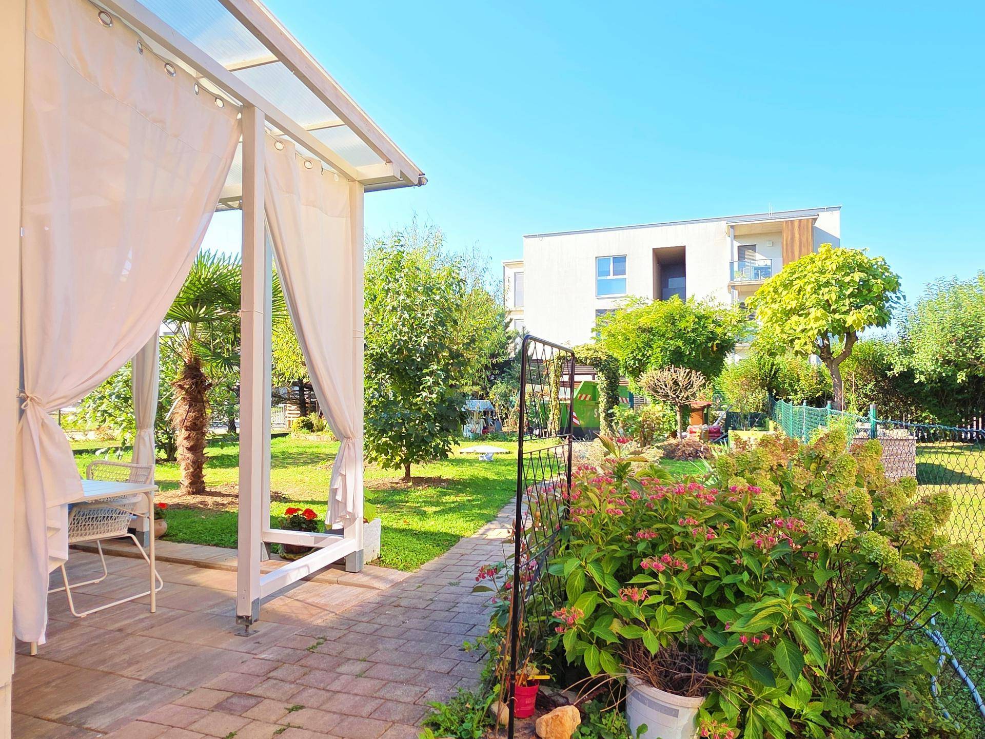 Gartenbereich mit überdachter Terrasse, Blumenbeeten und Blick auf ein Mehrfamilienhaus.