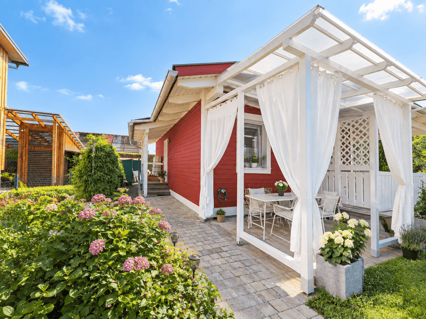Rotes Haus mit überdachter Terrasse, Gartenmöbeln und gepflegter Bepflanzung.