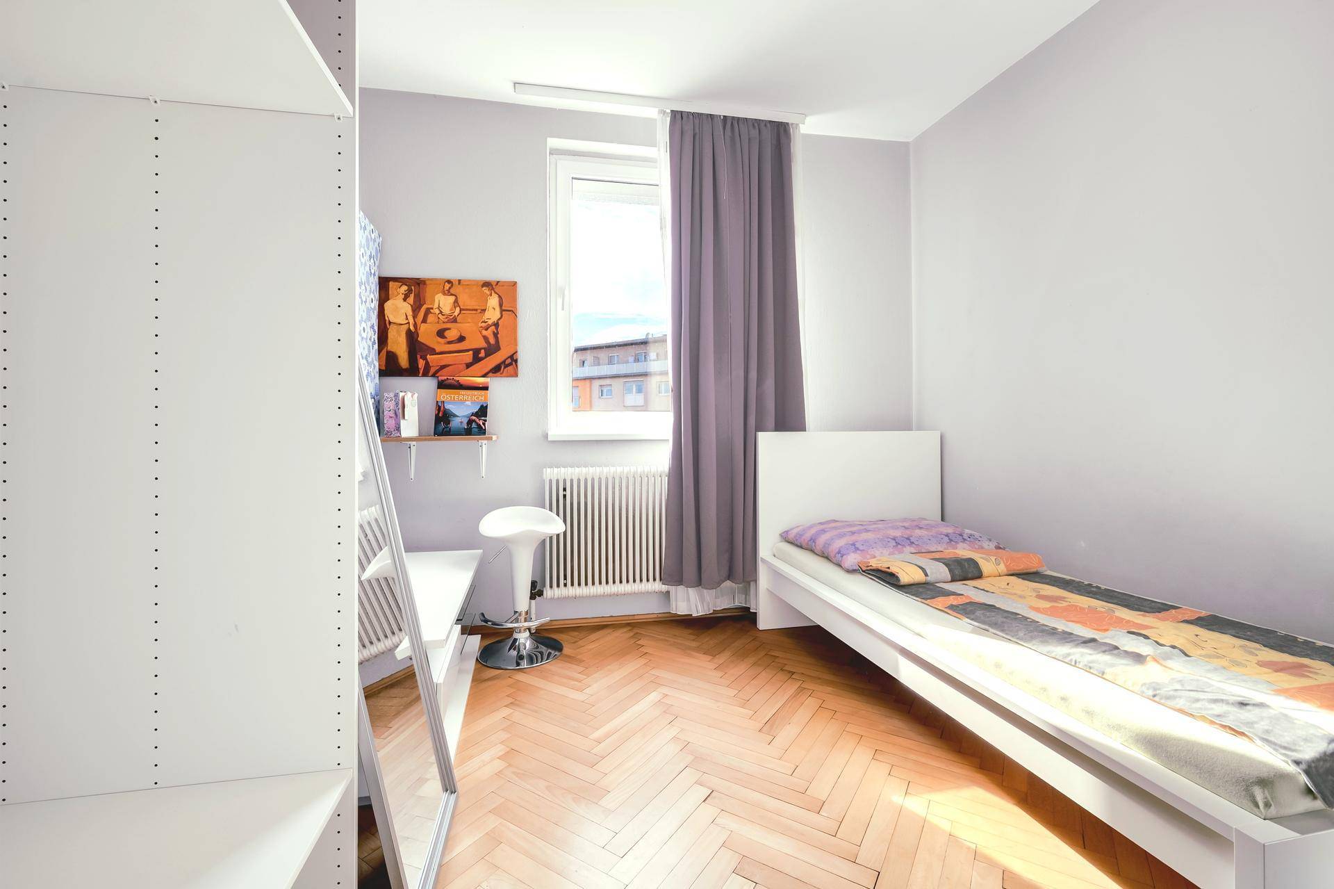 Helles Schlafzimmer mit Einzelbett, Parkettboden und großem Fenster.
