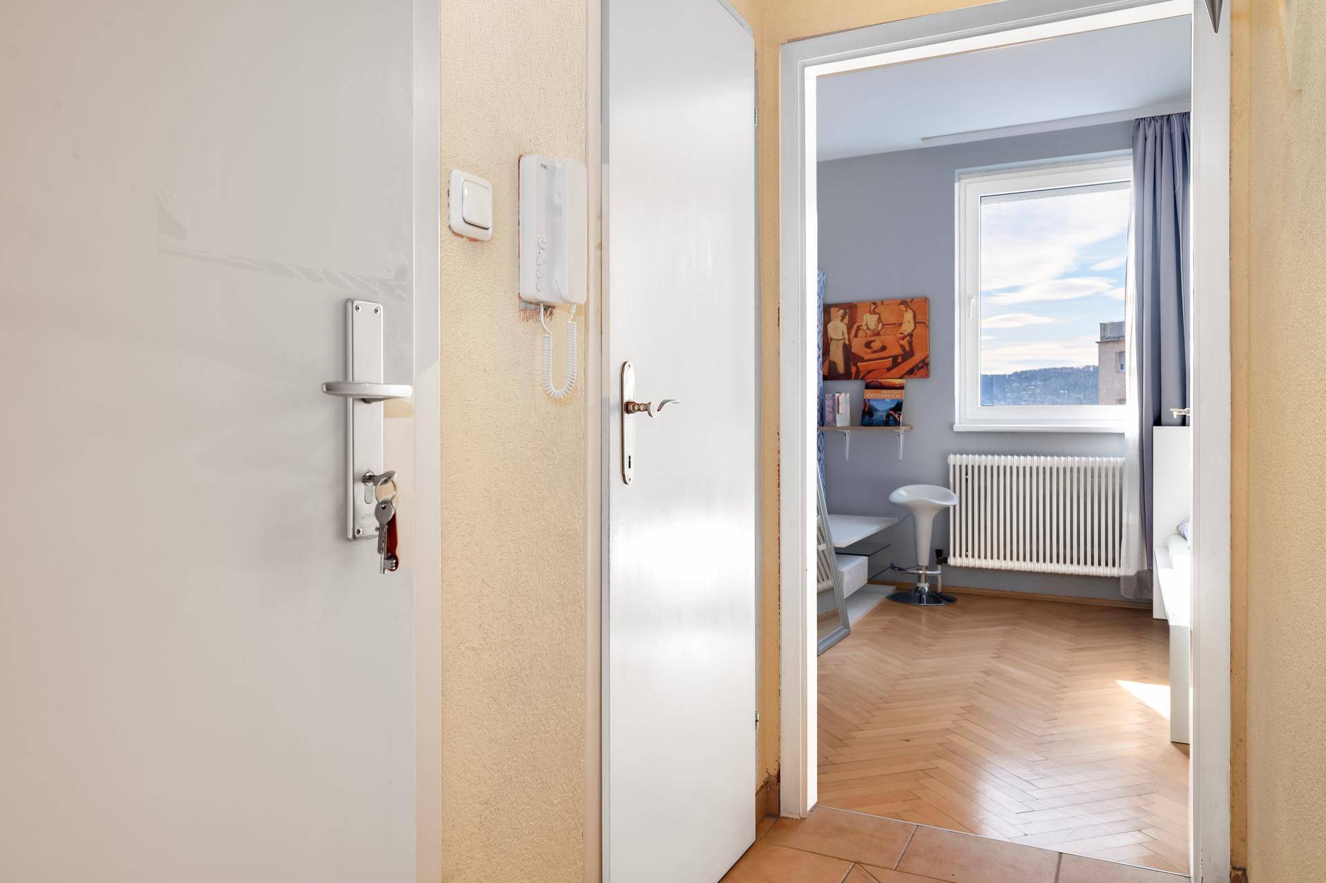 Flur mit Gegensprechanlage und Blick in ein helles Schlafzimmer mit Fenster.
