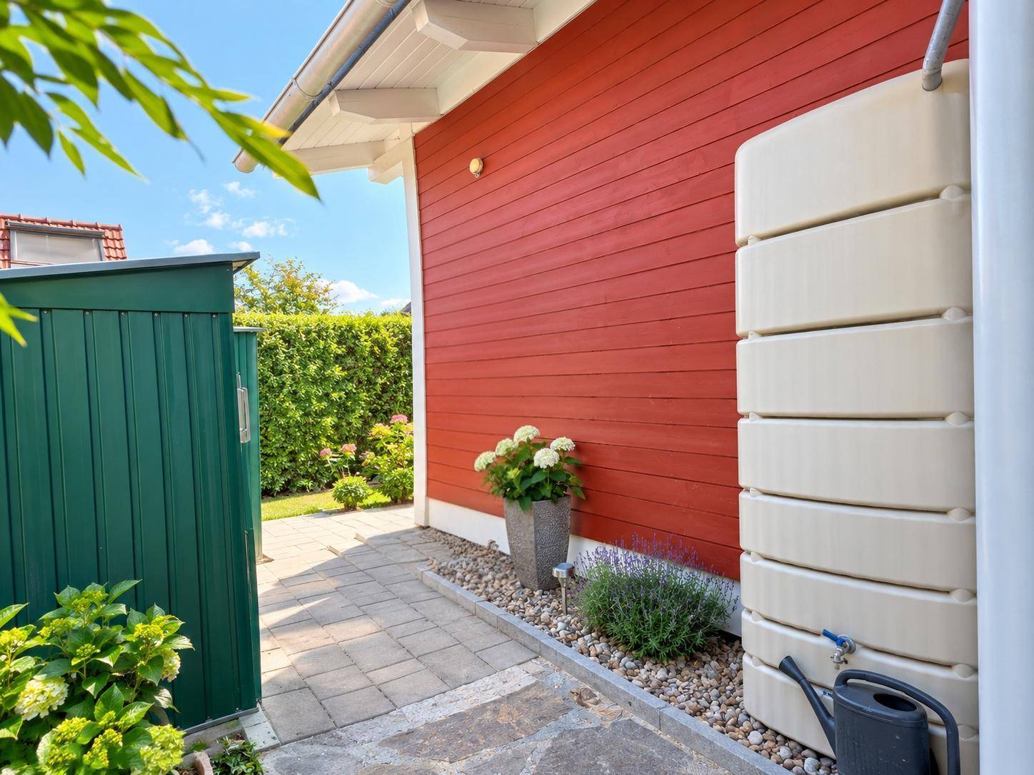 Rote Hauswand mit Regenwassertonne, grünem Gartenhaus und gepflastertem Zugangsbereich.