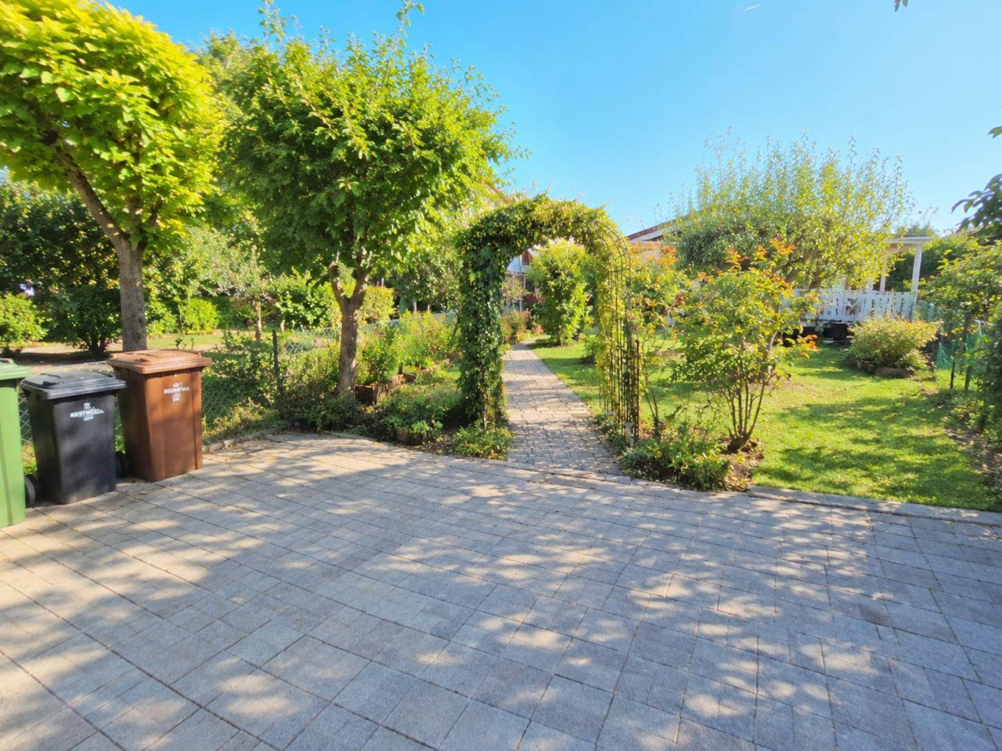 Gepflasterter Vorplatz mit Mülltonnen und einem grünen Torbogen zum Garten.