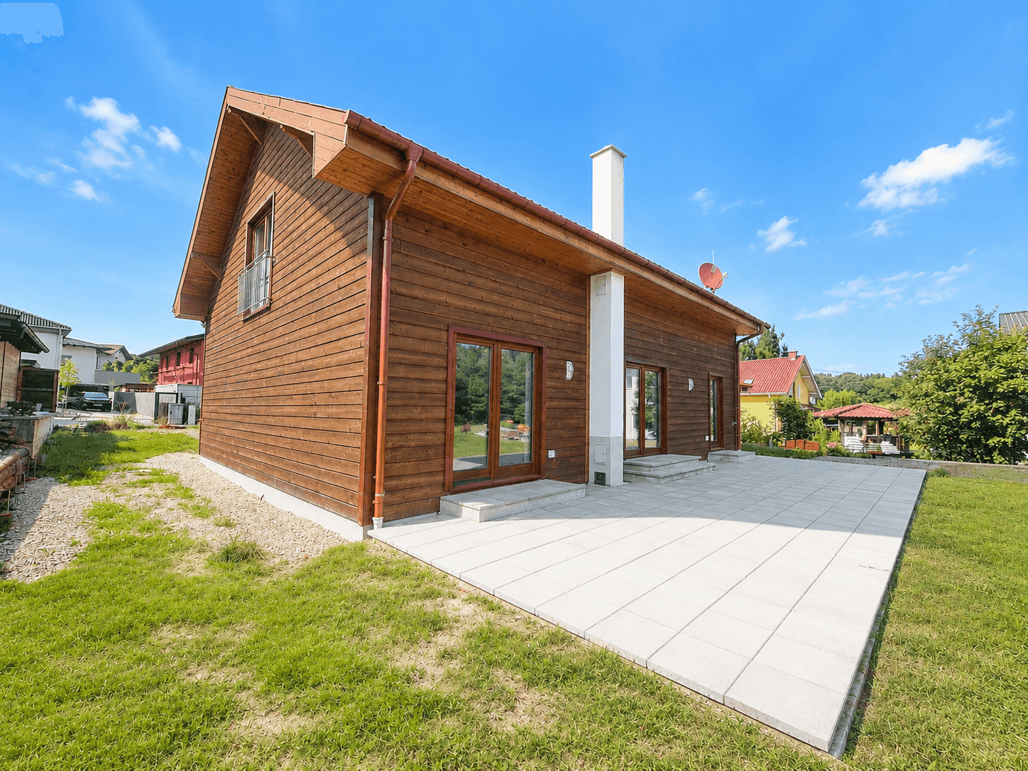 Rückseite des Holzhauses mit großer gepflasterter Terrasse und weitläufigem Rasenbereich.