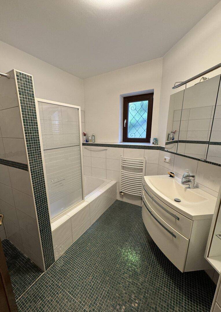 Badezimmer mit Dusche, Badewanne, Fenster und modernisierten Fliesen in Weiß und Grün.