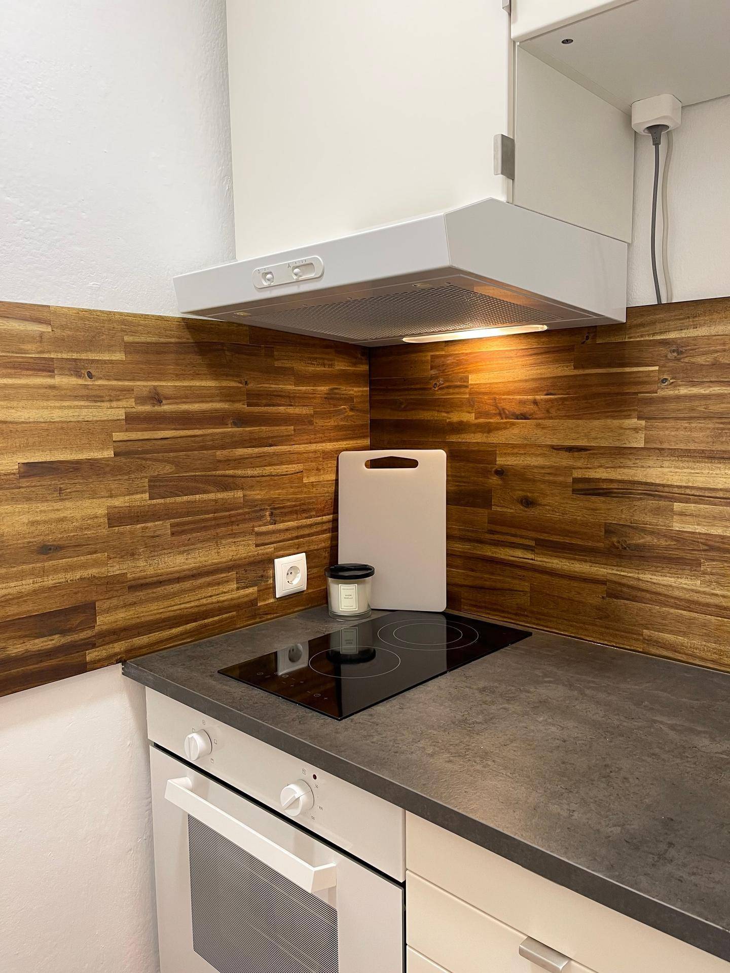 Kompakte Küchenzeile mit Induktionskochfeld, Backofen, Dunstabzugshaube und Holzrückwand.