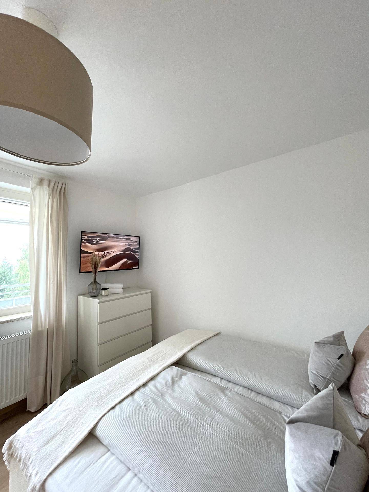 Schlafzimmer mit einem Flachbildfernseher über einer Kommode und einem Fenster mit hellen Vorhängen.