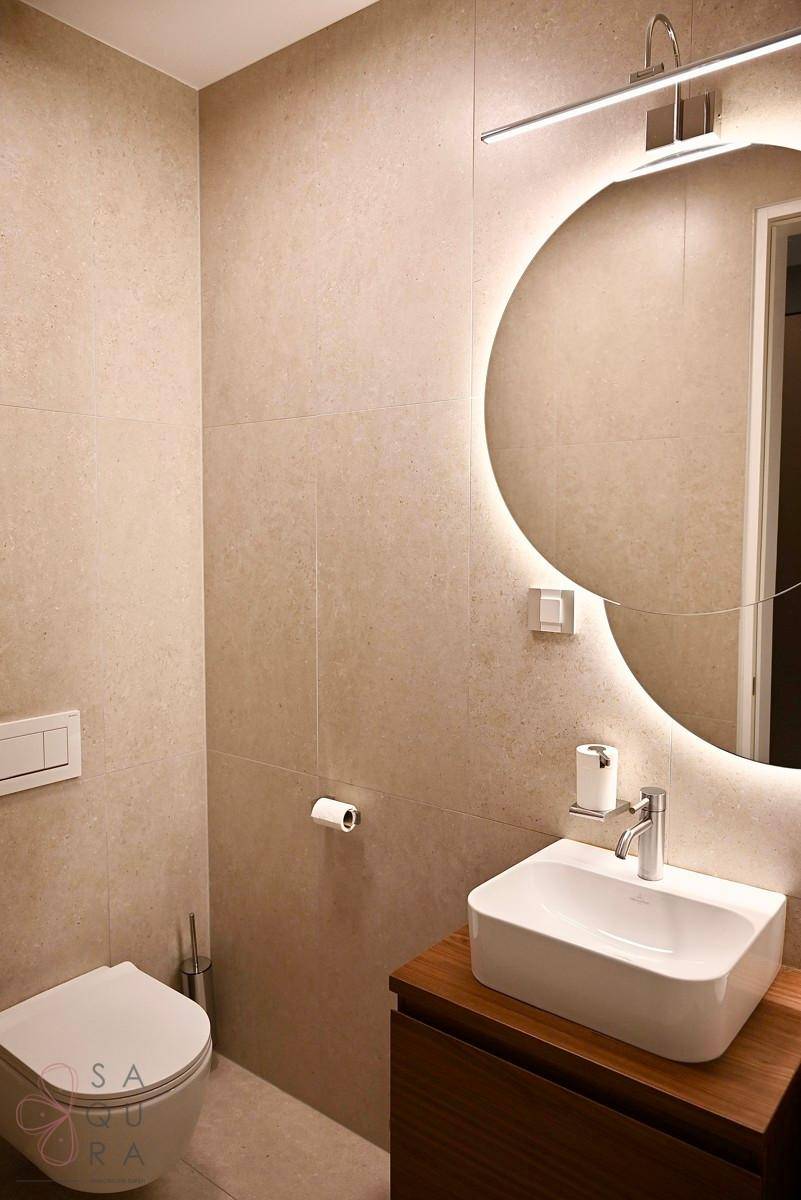 Modernes Gäste-WC mit beigen Fliesen, wandhängendem WC und beleuchtetem Rundspiegel.