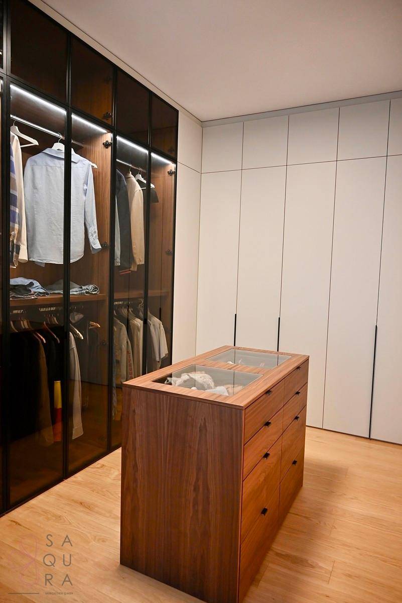 Begehbarer Kleiderschrank mit eleganten Glasfronten und einer zentralen Kommode aus Holz.