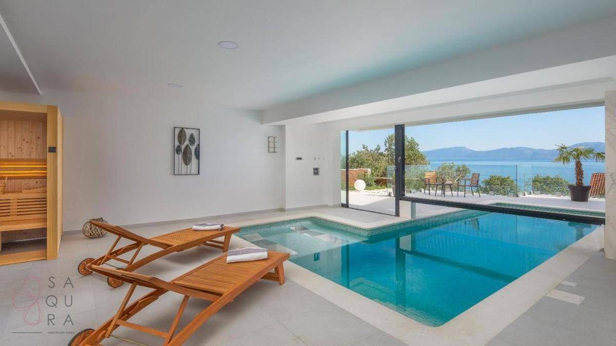 Heller Innenpoolbereich mit Sonnenliegen, Sauna und großen Schiebetüren zum Außenbereich mit Meerblick.