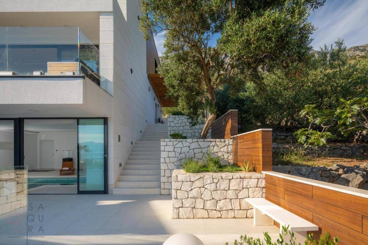 Seitlicher Blick auf die moderne Villa mit Steintreppen, Natursteinmauern und mediterraner Bepflanzung.