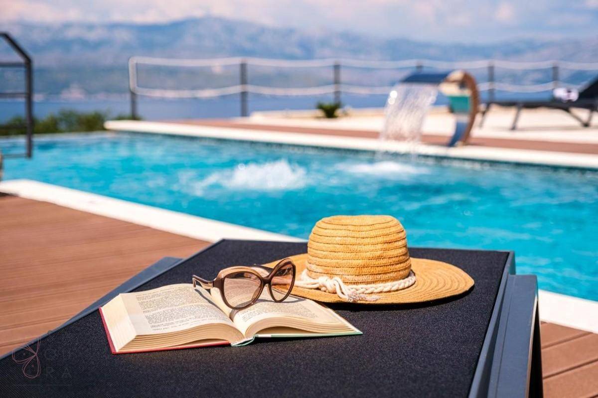 Sonnenliege am Pool mit Buch und Sonnenbrille, im Hintergrund das Meer und die Berge.