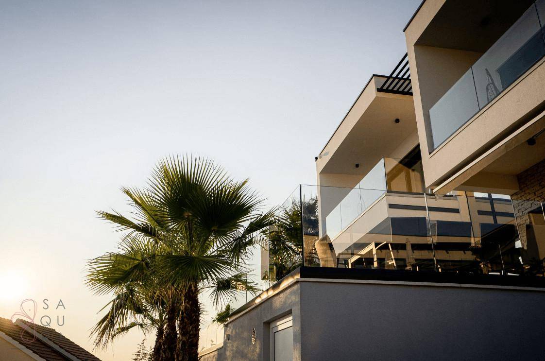 Detailansicht der modernen Villa mit Glasgeländern und Palmen vor hellem Himmel.