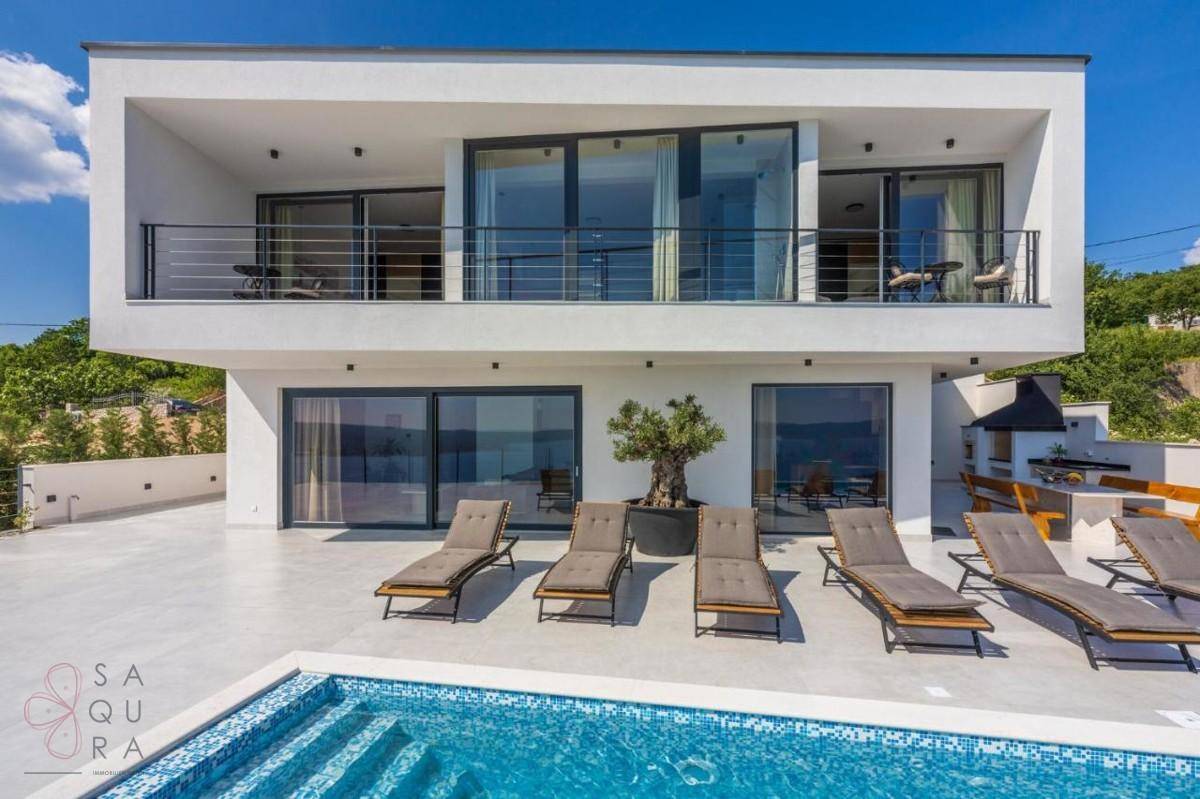 Moderne Villa mit Pool, Sonnenliegen und Balkonen, umgeben von grüner Landschaft unter blauem Himmel.