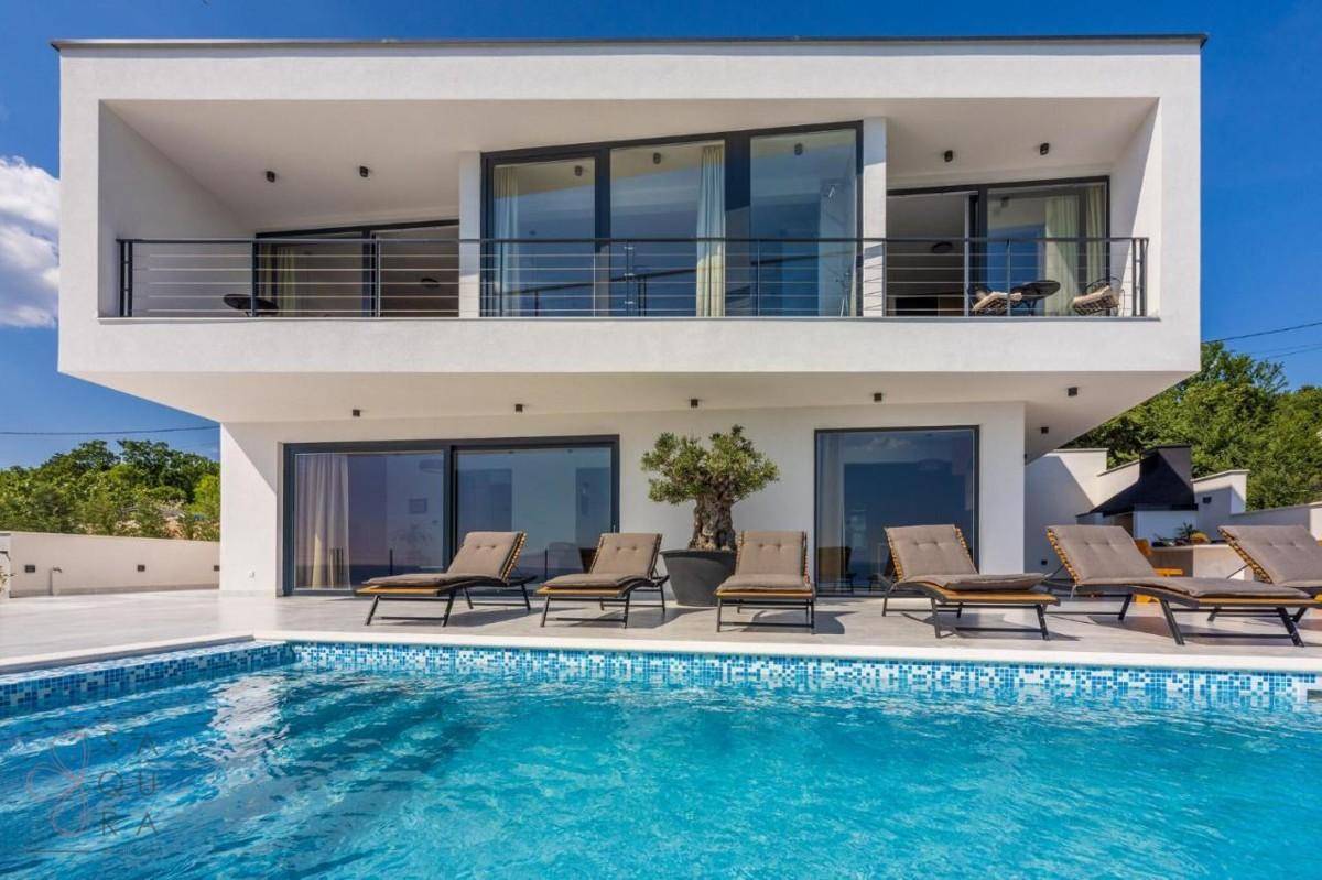 Moderne Villa mit Pool, Sonnenliegen und Balkonen, umgeben von grüner Landschaft unter blauem Himmel.