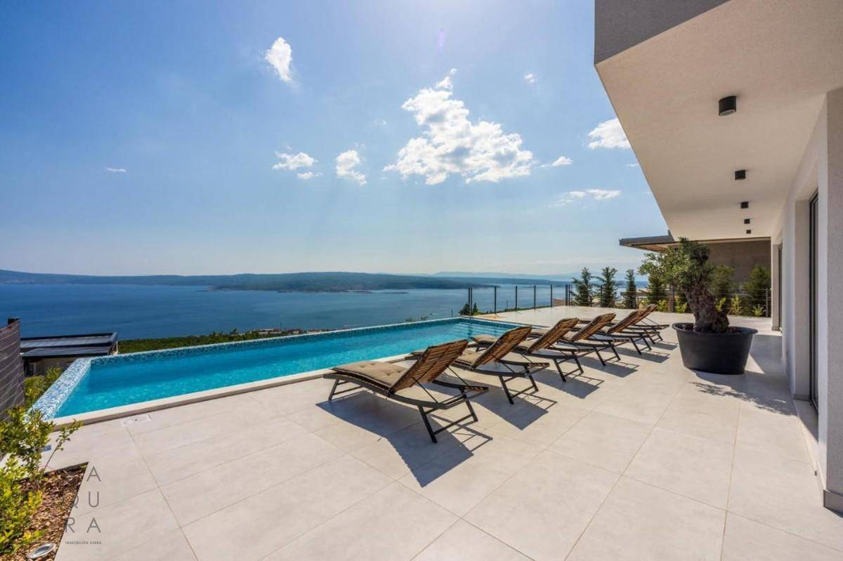 Weitläufiger Poolbereich mit Sonnenliegen und Panoramablick auf das glitzernde Meer und die Küste.