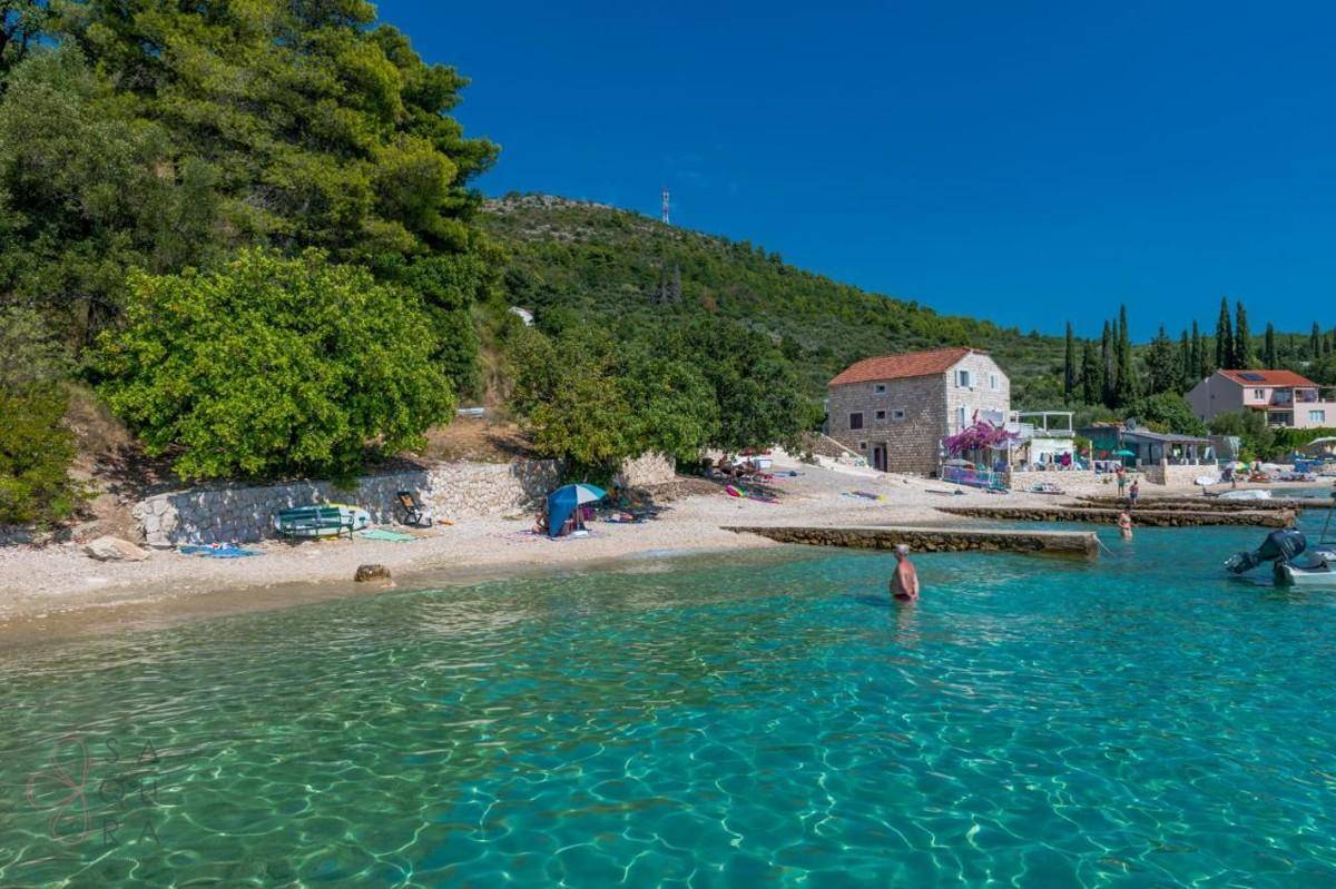 Malerischer Strand mit kristallklarem Wasser, umgeben von grünen Hügeln und einem traditionellen Gebäude.