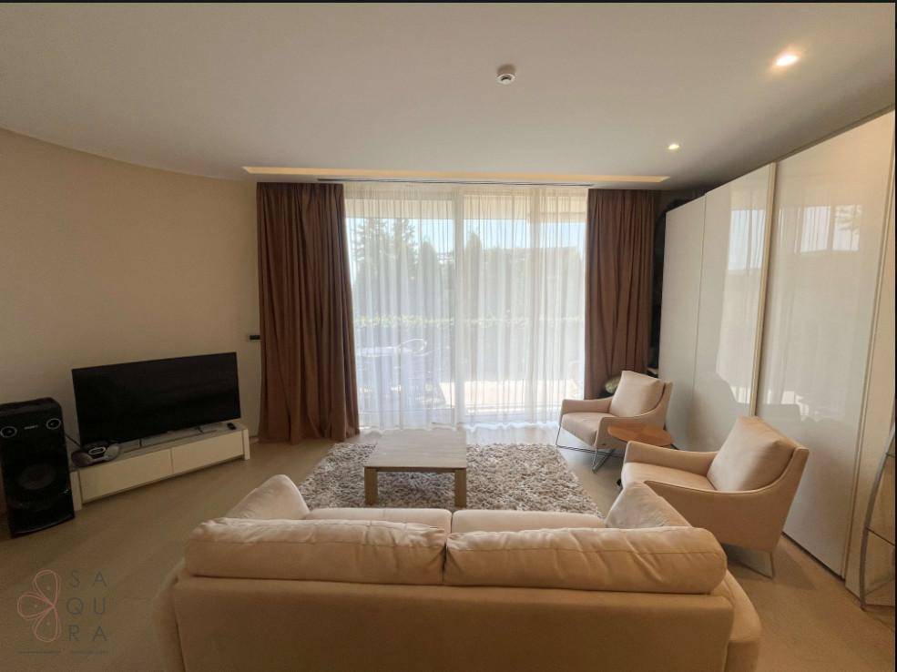 Gemütliches Wohnzimmer mit hellem Sofa, Teppich und großen Fenstern mit Vorhängen.