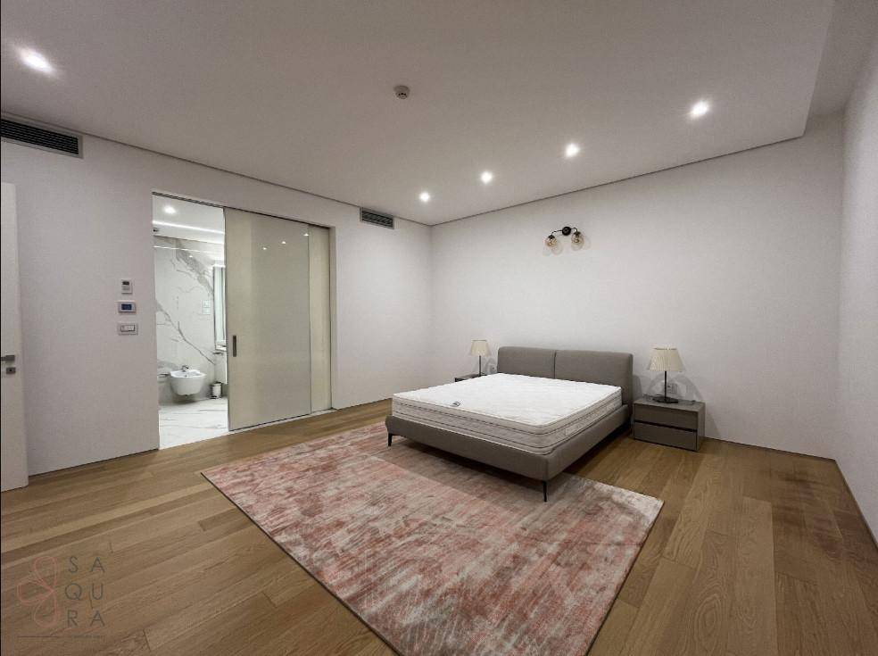 Schlafzimmer mit Doppelbett, Holzboden und direktem Zugang zu einem modernen Badezimmer durch eine Glasschiebetür.