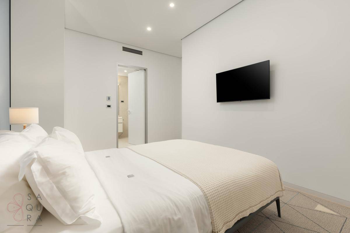 Ein minimalistisches Schlafzimmer mit Doppelbett, Wandfernseher und Zugang zu einem modernen Badezimmer.