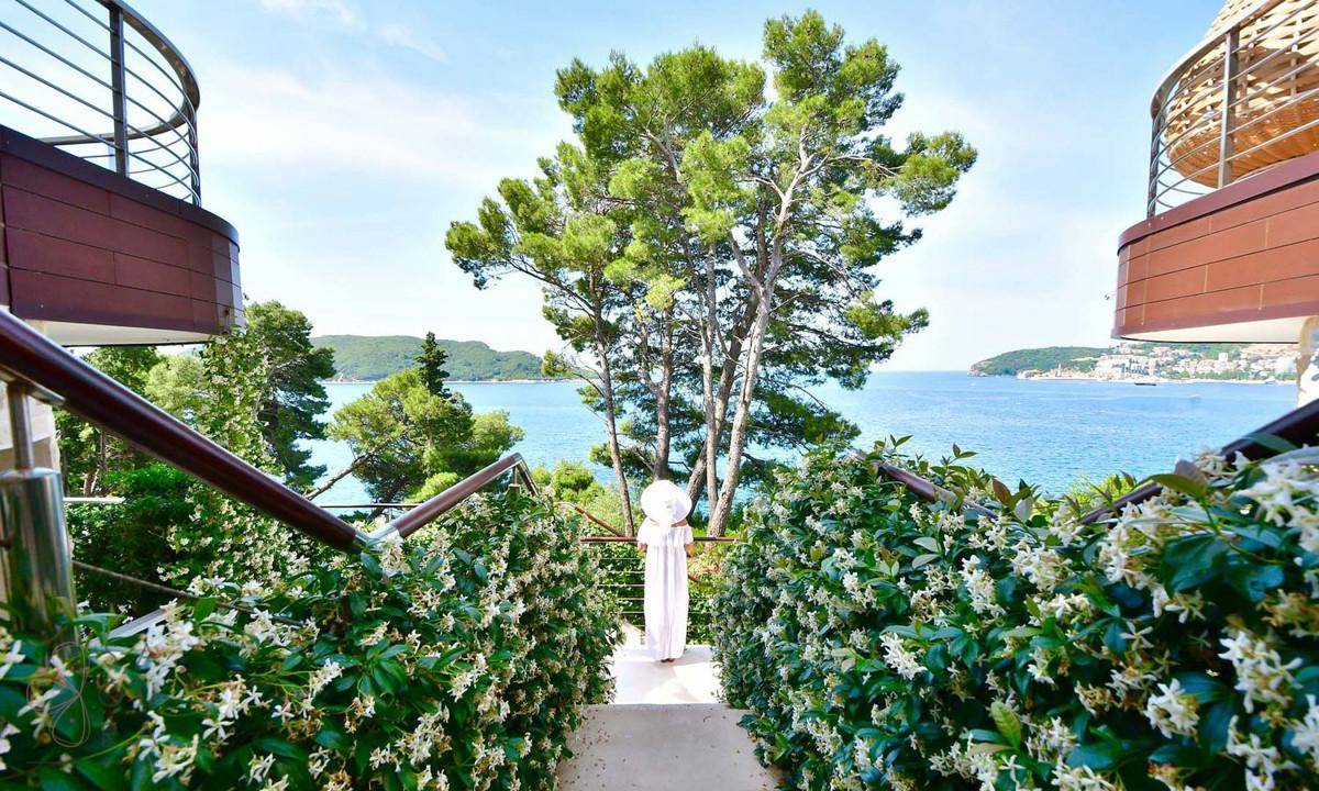 Eine Person steht auf einer Treppe, umgeben von blühenden Sträuchern, mit weitem Blick auf das Meer.