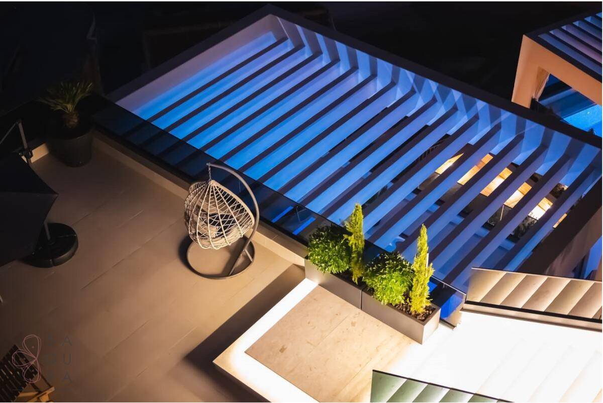 Dachterrasse bei Nacht mit beleuchteter Pergola, Sitzgelegenheiten und Pflanzen.
