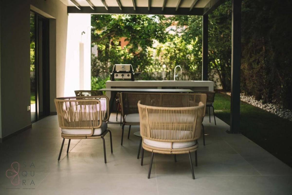 Gemütlicher Essbereich im Freien unter einer Pergola mit Grillstation im Hintergrund.