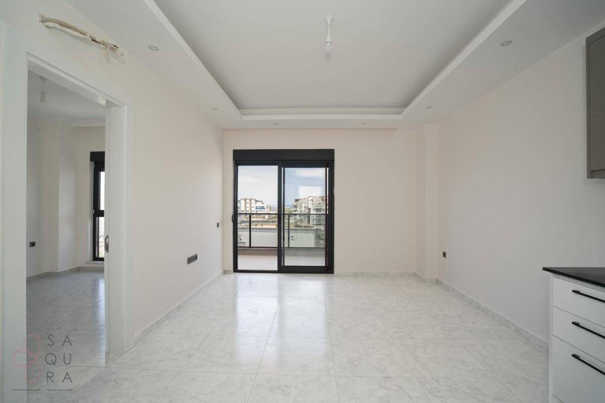 Heller, leerer Wohnbereich mit Fliesenboden und Zugang zu einem Balkon durch eine Schiebetür.