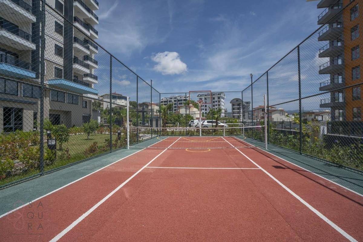 Roter Tennisplatz mit Netz und Zaun, umgeben von Grünflächen und Wohngebäuden.