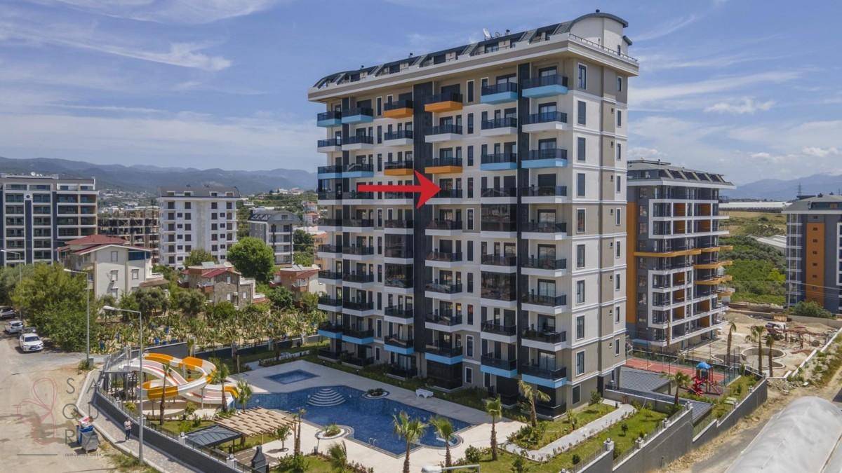 Luftaufnahme eines modernen Wohnkomplexes mit Pool, Wasserrutsche und Tennisplatz, umgeben von Grünflächen.