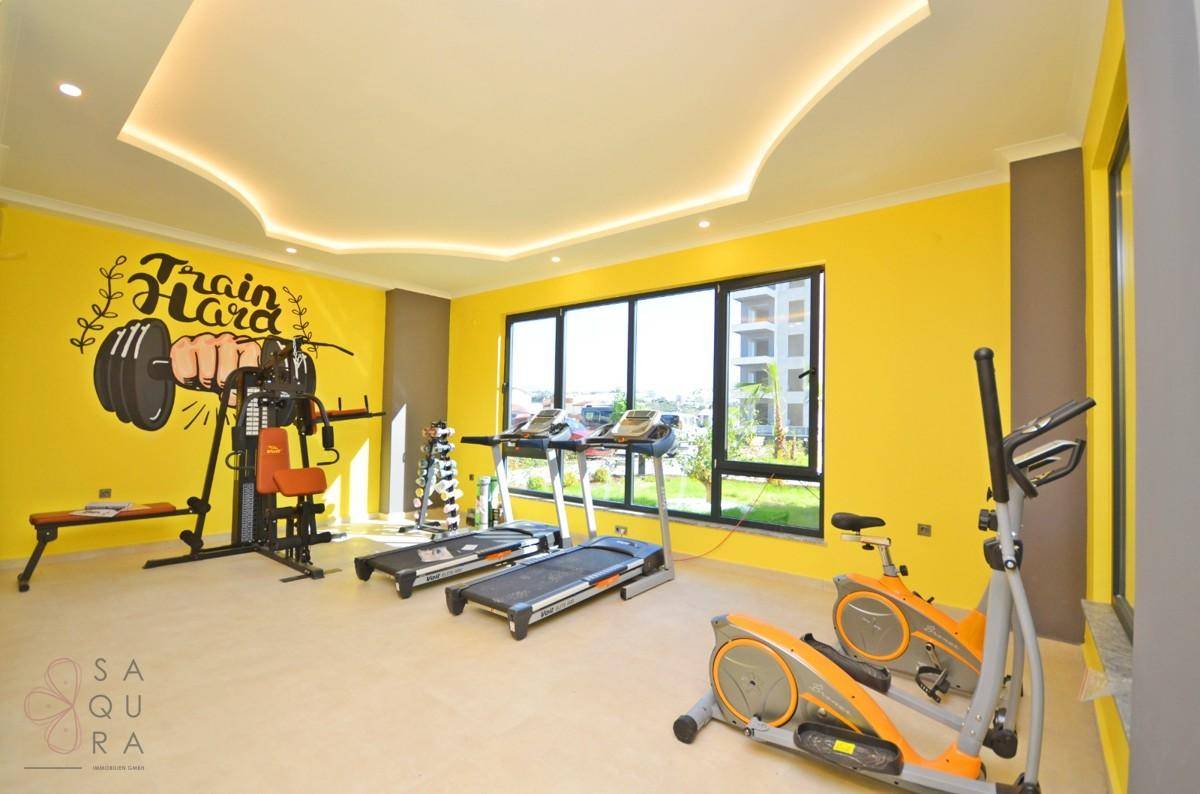Moderner Fitnessraum mit gelben Wänden, verschiedenen Trainingsgeräten und großen Fenstern für Tageslicht.