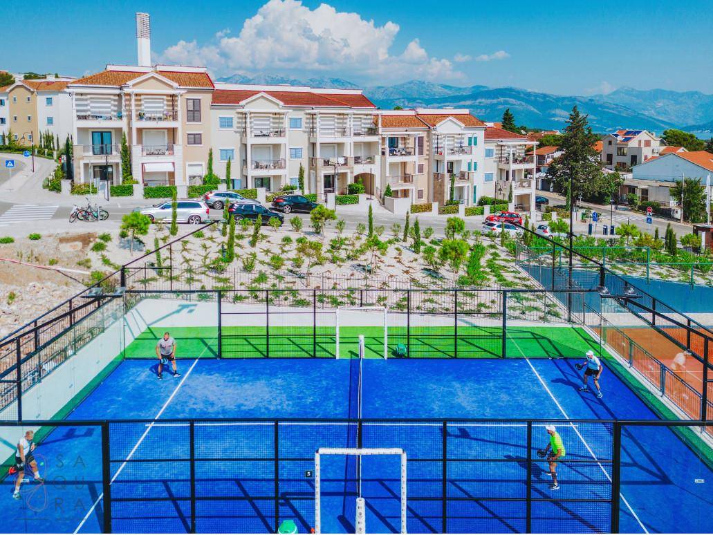 Padel-Tennisplatz mit Spielern vor modernen Wohngebäuden und Bergkulisse unter blauem Himmel.