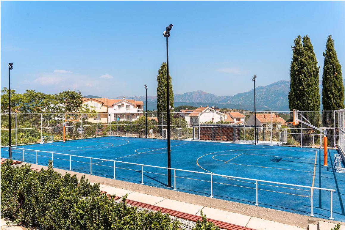 Blauer Basketballplatz im Freien mit Flutlichtmasten und Bergblick, umgeben von Wohngebäuden.