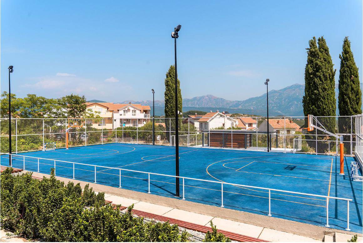 Blauer Basketballplatz mit Flutlichtmasten, umgeben von Bäumen und Häusern im Hintergrund.