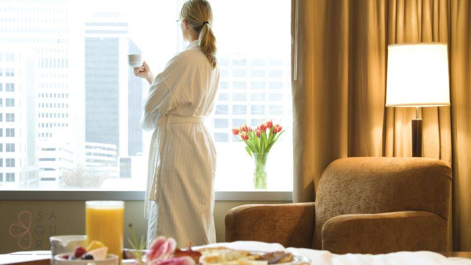 Frau im Bademantel genießt den Stadtblick aus einem hellen Schlafzimmer mit Frühstück.