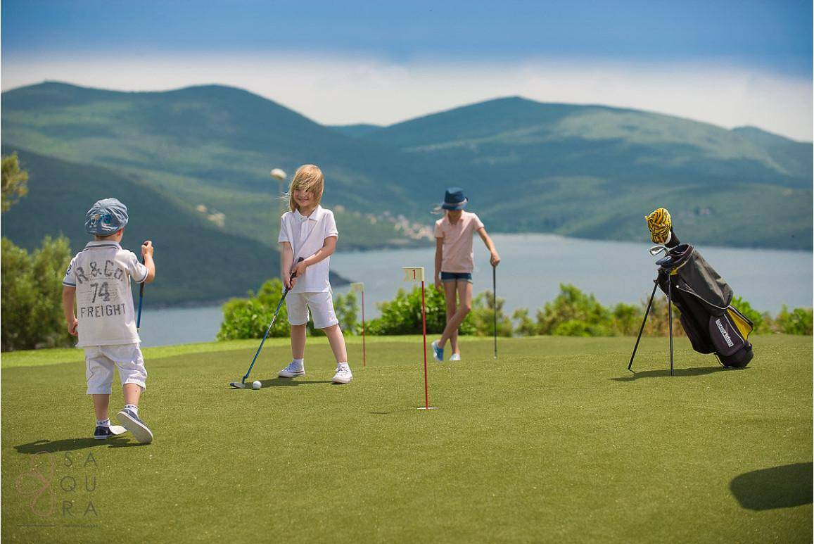 Kinder spielen Golf auf einem Putting Green mit Meer- und Bergblick im Hintergrund.