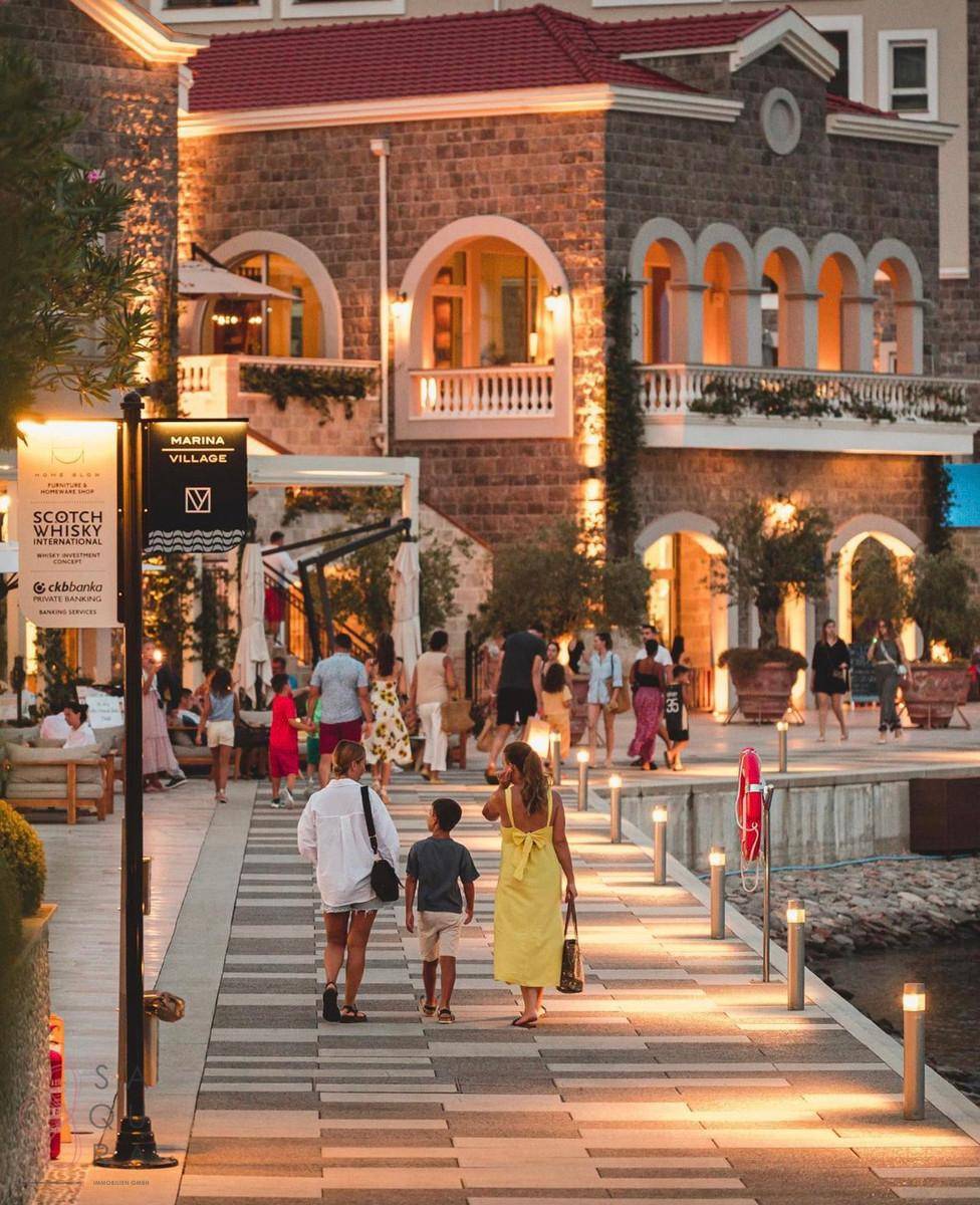 Lebhafte Promenade in einem Marina Village mit Fußgängern und beleuchteten Gebäuden.